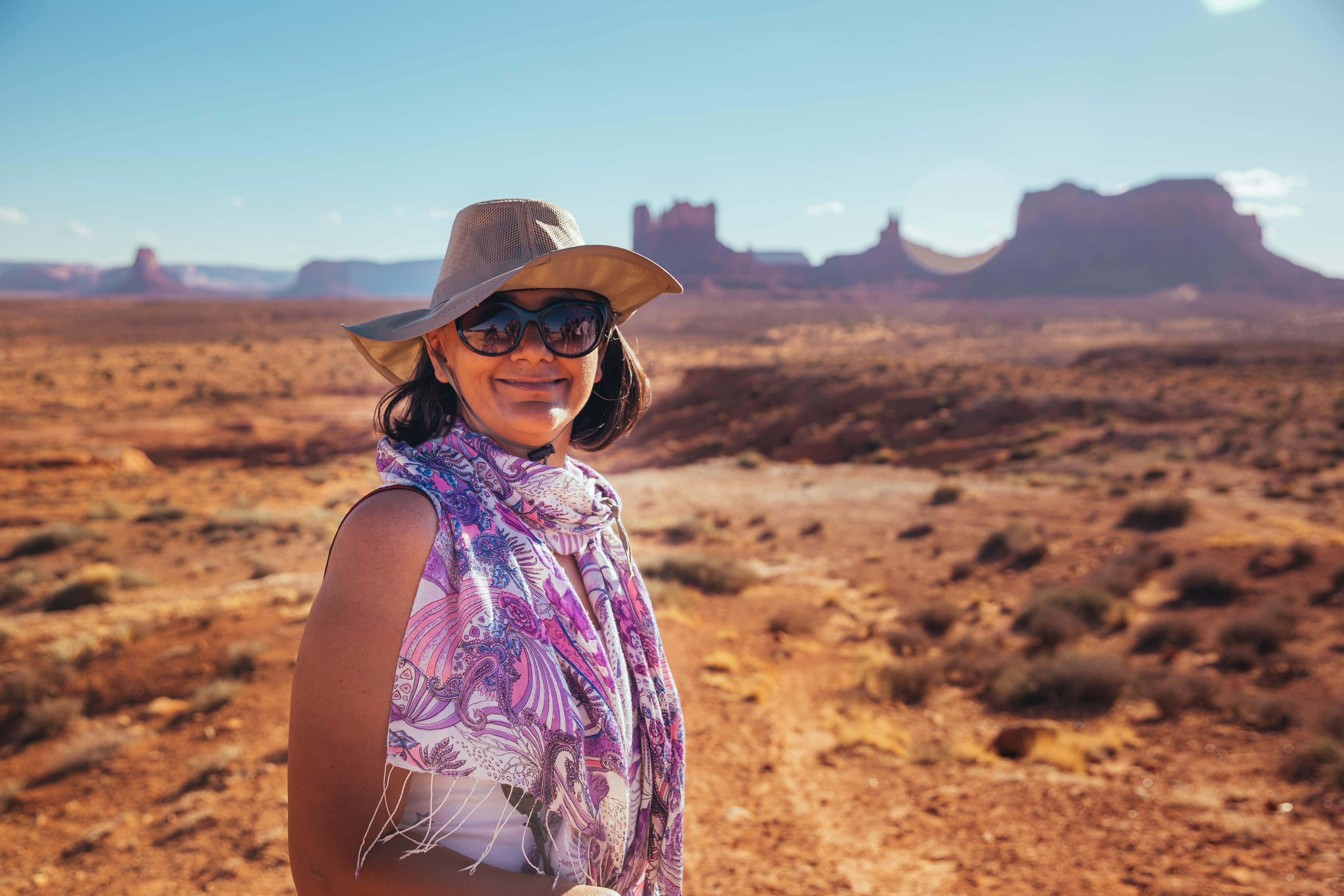 A woman wearing a scarf, sunglasses and a hat smiles with the backdrop of desert mesas in Monument Valley