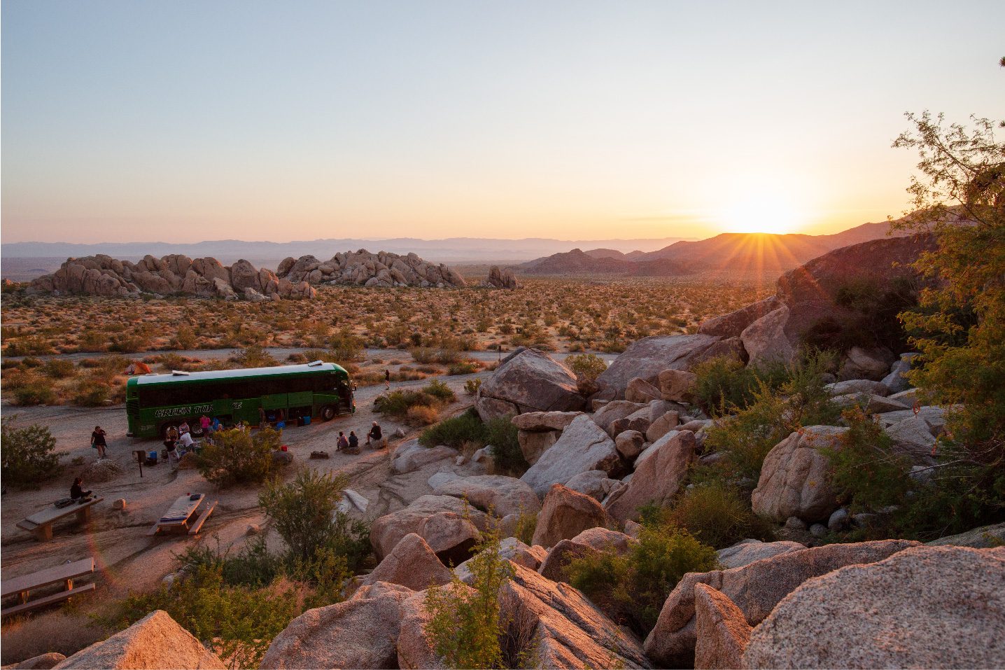 A group of explorers visiting Joshua Tree National park relax during a sunset. The mid-size group is lounging around the Green Tortoise adventure bus.