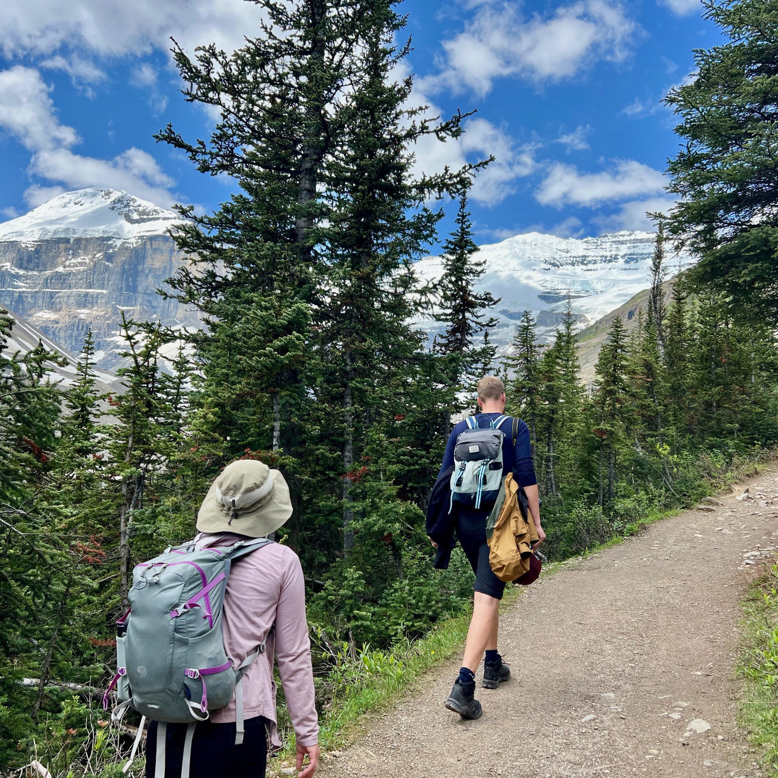 Two people hike with backpacks in the Canadian Rockies at Banff National Park. Trees line the dirt path with snow-covered mountains in the distance.