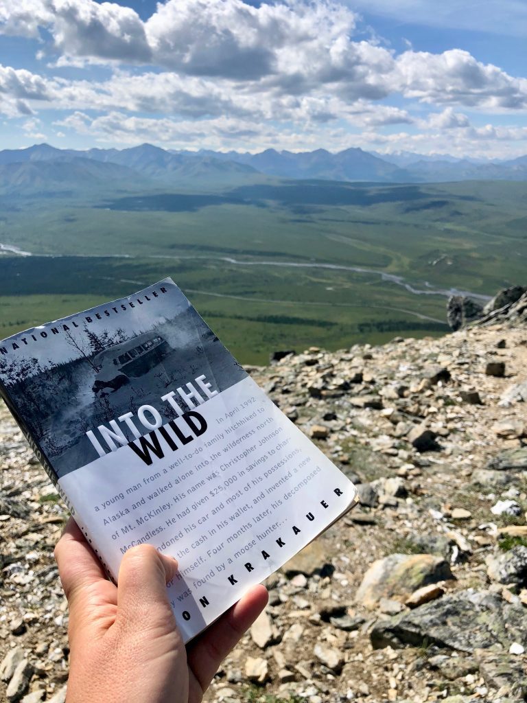 A person holds up a book titled "Into The Wild" by Jon Krakauer on a trail in Denali National Park
